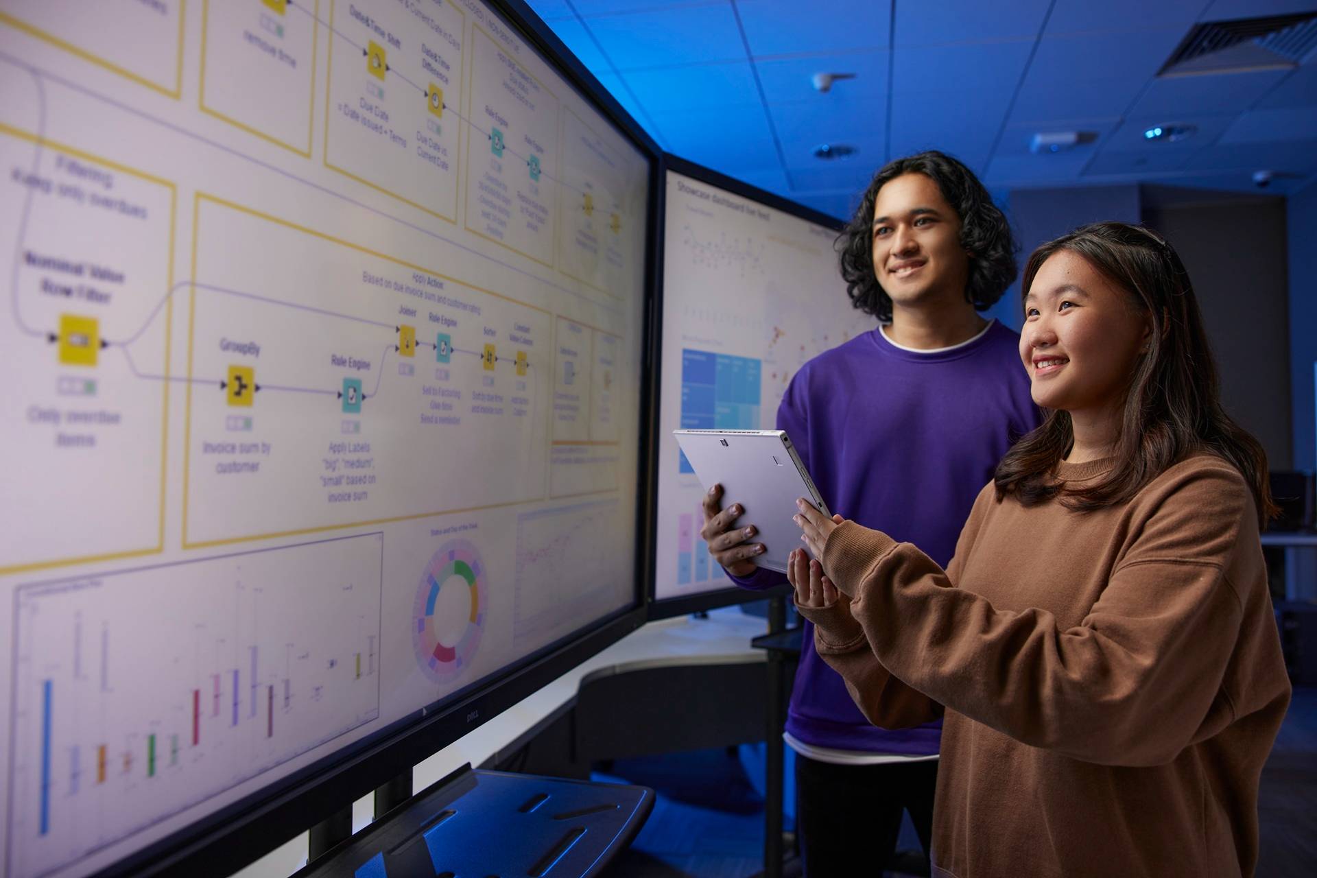 Two students reviewing data-analytics workflows on a large display screen, with one holding a tablet in a modern lab setting.