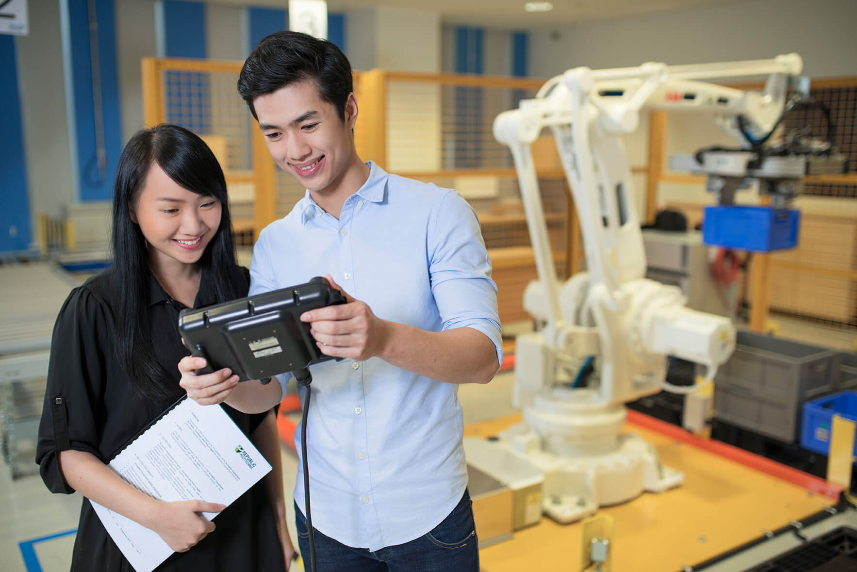 A pair of student engineers in a supply chain lab with a robotic arm in the background