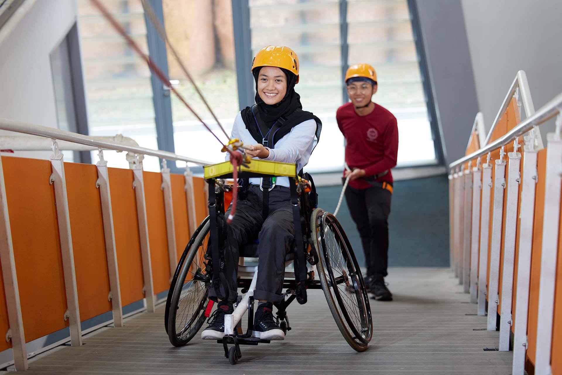 A happy student on wheelchair surmounting the xperiential learning centre on a ramp, with a fellow student as a belayer in the background