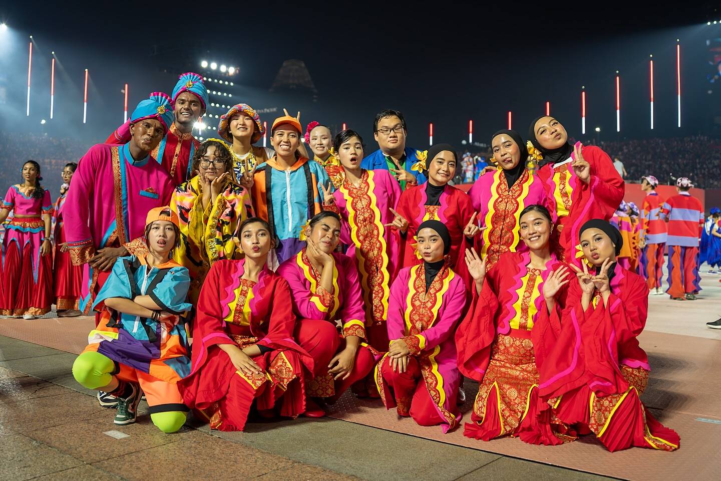 A large group of RP students dressed in colorful traditional and modern costumes posing joyfully at a National Day Parade event with bright stage lights and cheering crowds in the background.