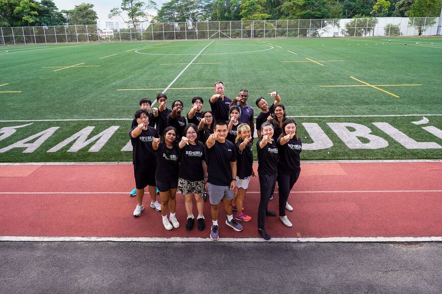 A diverse group of students standing together on a running track, smiling and pointing towards the camera with a sports field in the background.