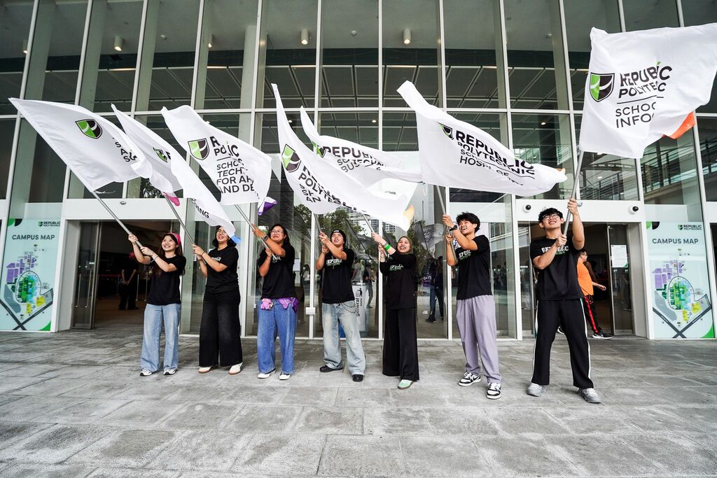 Students standing outside a campus building, waving Republic Polytechnic school flags and welcoming participants during an on-campus event.